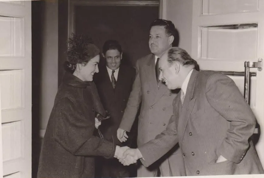 Archive photograph: a woman being greeted with a handshake at a doorway, with two men in suits looking on