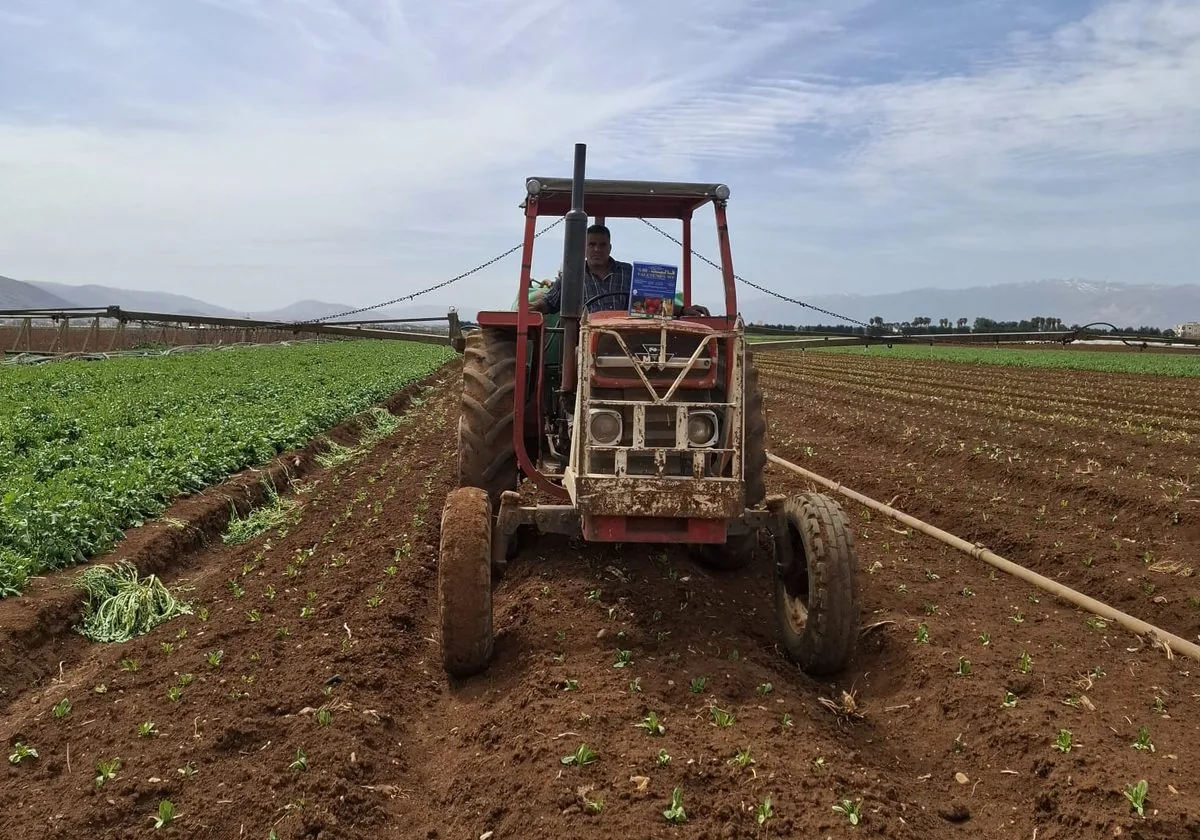 A tractor with a boom sprayer working a newly planted field, with a product pack on the bonnet and mountains behind